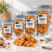 Load image into Gallery viewer, Four stand-up pouches of Sugar &amp; Salt Peanut Brittle in different sizes arranged on a marble surface, with several pieces of the brittle spilled in front. A small bowl of mixed nuts and a folded cloth are visible in a bright kitchen-like setting.