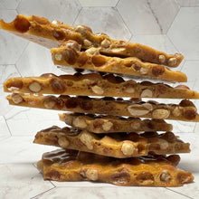 Load image into Gallery viewer, A stack of five golden-brown peanut brittle bark pieces with chopped peanuts, set against a light gray marble-patterned background.